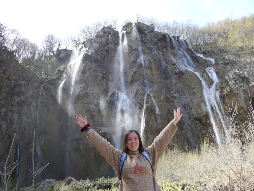 Tiani Travels standing in front of a big waterfall with her arms in the air at Plitvice Lakes, Croatia